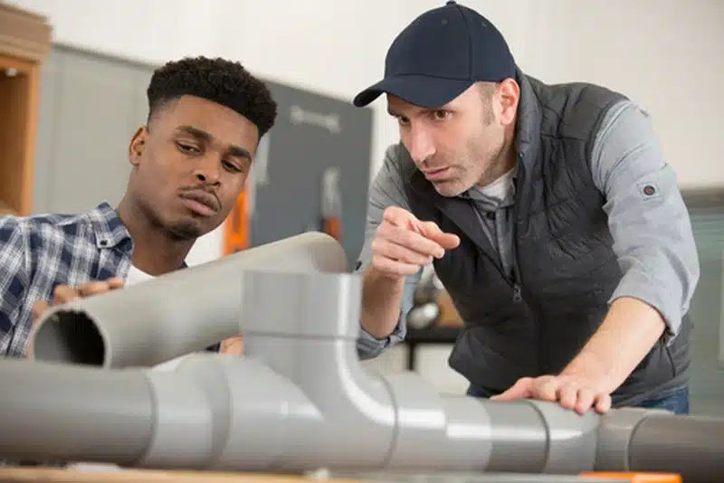 Two male plumbers working together to examine several gray PVC pipe fittings on a workbench.