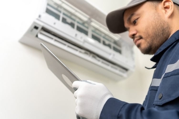 An HVAC technician wearing a cap and gloves looks down at a tablet while inspecting a white, wall-mounted mini-split air conditioner.