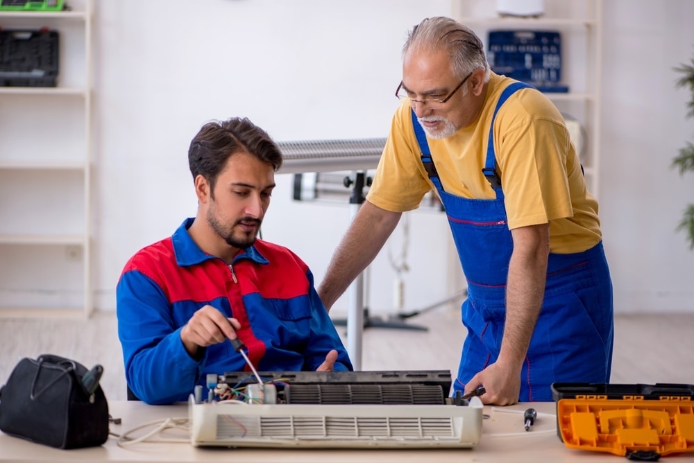 Two technicians in blue overalls repairing a disassembled indoor air conditioner unit at a workshop table.