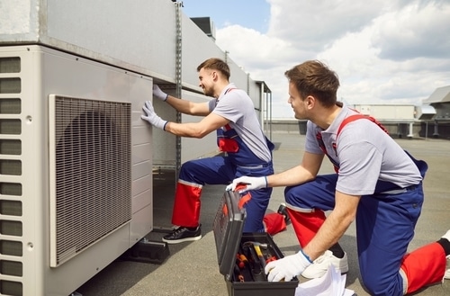 Two technicians in blue and red overalls repair a commercial rooftop HVAC air conditioning unit.
